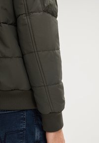 Giacca a vento verde con orlo e polsini a coste, caratterizzata da una texture trapuntata e una vestibilità slim; dettaglio della cucitura visibile sulla manica.