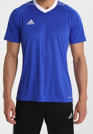 Mann trägt ein blaues Adidas kurzärmeliges Sportshirt mit weißem Kragen und schwarzen Adidas-Shorts, steht mit entspannten Armen.