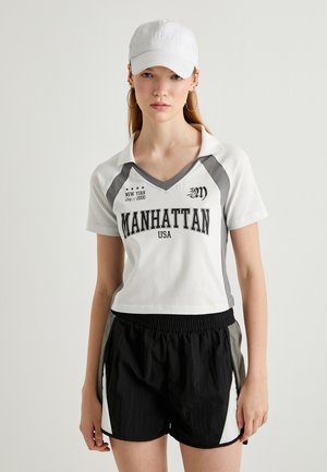 Hvid polo-shirt med grå detaljer, "MANHATTAN USA" tryk, parret med sorte shorts med hvide og grå sidepaneler. Hvid kasket.
