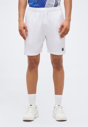 Άνδρας που φοράει λευκό αθλητικό σορτς με μαύρο λογότυπο ασπίδας στο αριστερό πόδι, λευκές κάλτσες, λευκά αθλητικά παπούτσια και μπλε πουκάμισο με σχέδια.