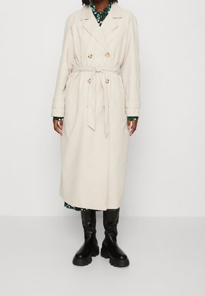 Persona con gabardina beige de doble botonadura con cinturón, vestido floral debajo y botas negras gruesas hasta la rodilla, de pie sobre fondo blanco.