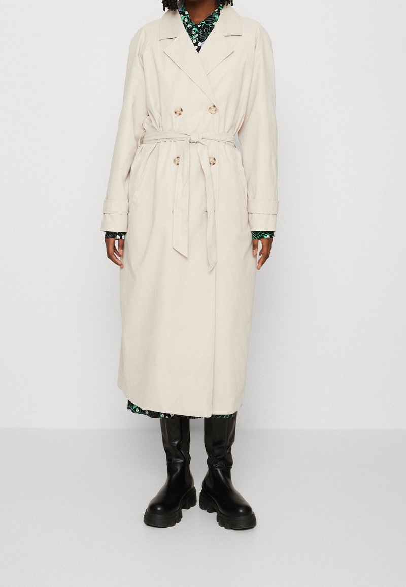 Personne portant un trench-coat beige croisé avec ceinture, robe fleurie en dessous, et bottes noires épaisses montant jusqu'aux genoux, debout devant un fond blanc.