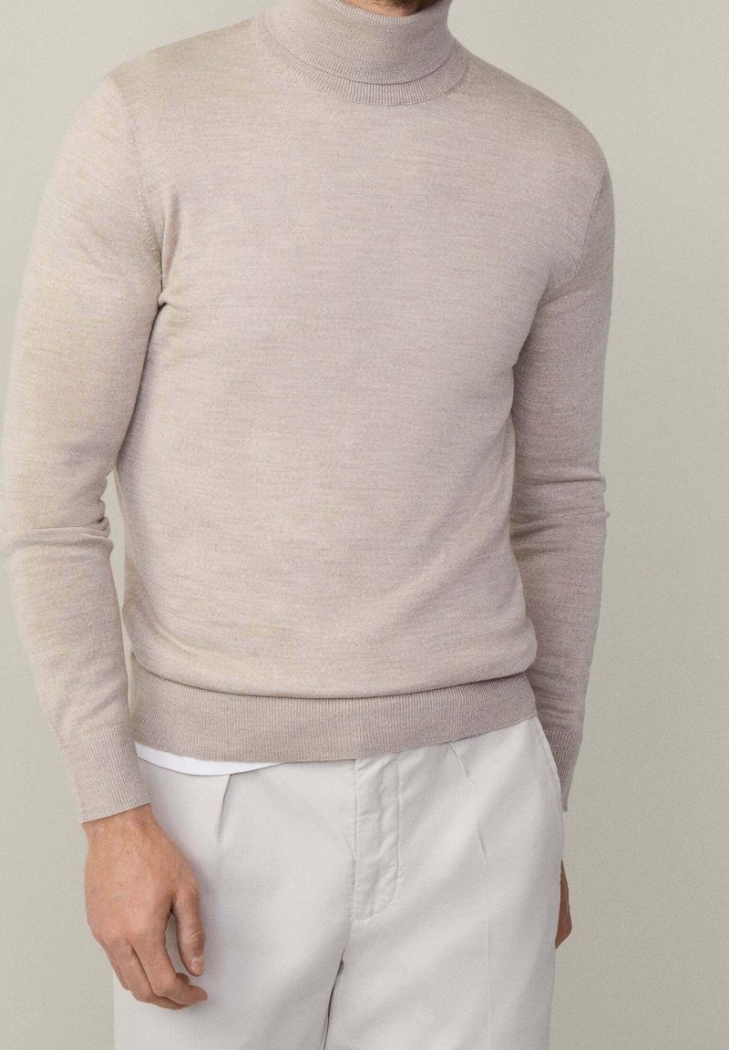 Homme portant un pull à col roulé beige et un pantalon blanc devant un fond gris clair uni, avec une main détendue sur le côté.
