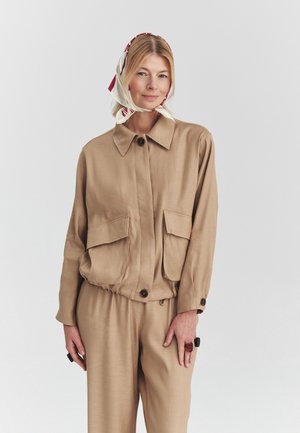 Femme portant une veste beige boutonnée et un pantalon, avec un foulard crème et rouge à motifs, debout devant un fond clair uni.