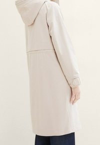 Abrigo largo de color beige claro con capucha, puños abotonados y cintura ajustada. Textura suave con un diseño sencillo y minimalista.