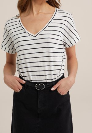 Vrouw draagt een wit V-hals shirt met zwarte horizontale strepen en een zwarte rok met een dubbele ring riem, handen in de zakken.