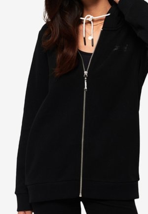 Kobieta ubrana w czarną kurtkę na zamek na czarny top z białym sznurkiem zawiązanym w kokardę wokół szyi.