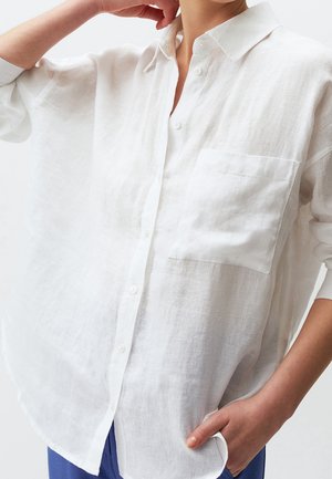 Personne portant une chemise blanche ample boutonnée avec une poche poitrine et un pantalon bleu, une main dans la poche, l’autre près du cou.