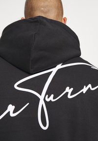 Sudadera negra con un gran texto en estilo de escritura a mano en blanco en la parte trasera. El material parece suave con una textura lisa y un diseño de capucha clásico.