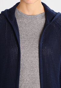 Granatowy dzianinowy kardigan z teksturowanym wzorem, z kapturem i otwartym przodem, noszony na szarej koszulce. Skoncentrowano się na oddychających, lekkich materiałach.