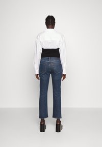 Camisa blanca de manga larga con un tejido negro recortado encima, combinada con jeans rectos azules que presentan un detalle en el bolsillo trasero y tacones de plataforma negros.