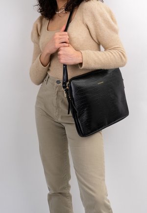 Zwarte handtas van leer met krokodillenstructuur in een rechthoekige vorm, voorzien van een gouden logo en een afneembare riem. Vrouwelijk figuur die de tas vasthoudt.