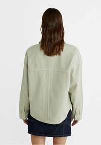 Kobieta z długimi brązowymi włosami nosi oversize'ową jasnoskalistą kurtkę i ciemnoniebieską spódnicę dżinsową, widoczna od tyłu na białym tle.