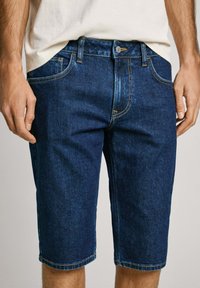 Donkere spijkershorts met een rechte snit, vijf zakken, gele stiksels en een knoopsluiting met een metalen klinknagel.