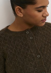 Brun, texturerad kofta med rund halsringning och två svarta knappar. Tyget verkar vara ulligt och kraftigt, med en naturlig look.