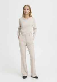 Haut en tricot beige à manches longues avec un col rond, associé à un pantalon taille haute beige clair, ajusté. Chaussures noires et mains dans les poches.