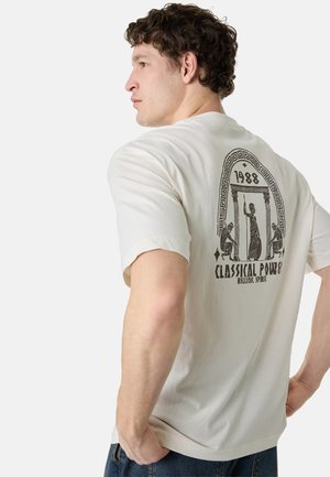 Mann in weißem T-Shirt mit schwarzem klassischem griechischem Design, Säulen und Figuren, Text "1988 Classical Power Hellenic Spirit" auf dem Rücken, seitlich blickend.