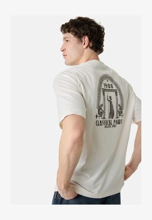 Uomo con maglietta bianca con design classico greco nero, colonne e figure, testo "1988 Classical Power Hellenic Spirit" sulla schiena, rivolto di lato.