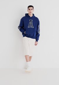 Námořnický modrý sweatshirt s velkým bílým potiskem "A", navržený s kapucí na šňůrky. V kombinaci s krémovými šortkami a bílými teniskami. Minimalistický celkový vzhled.