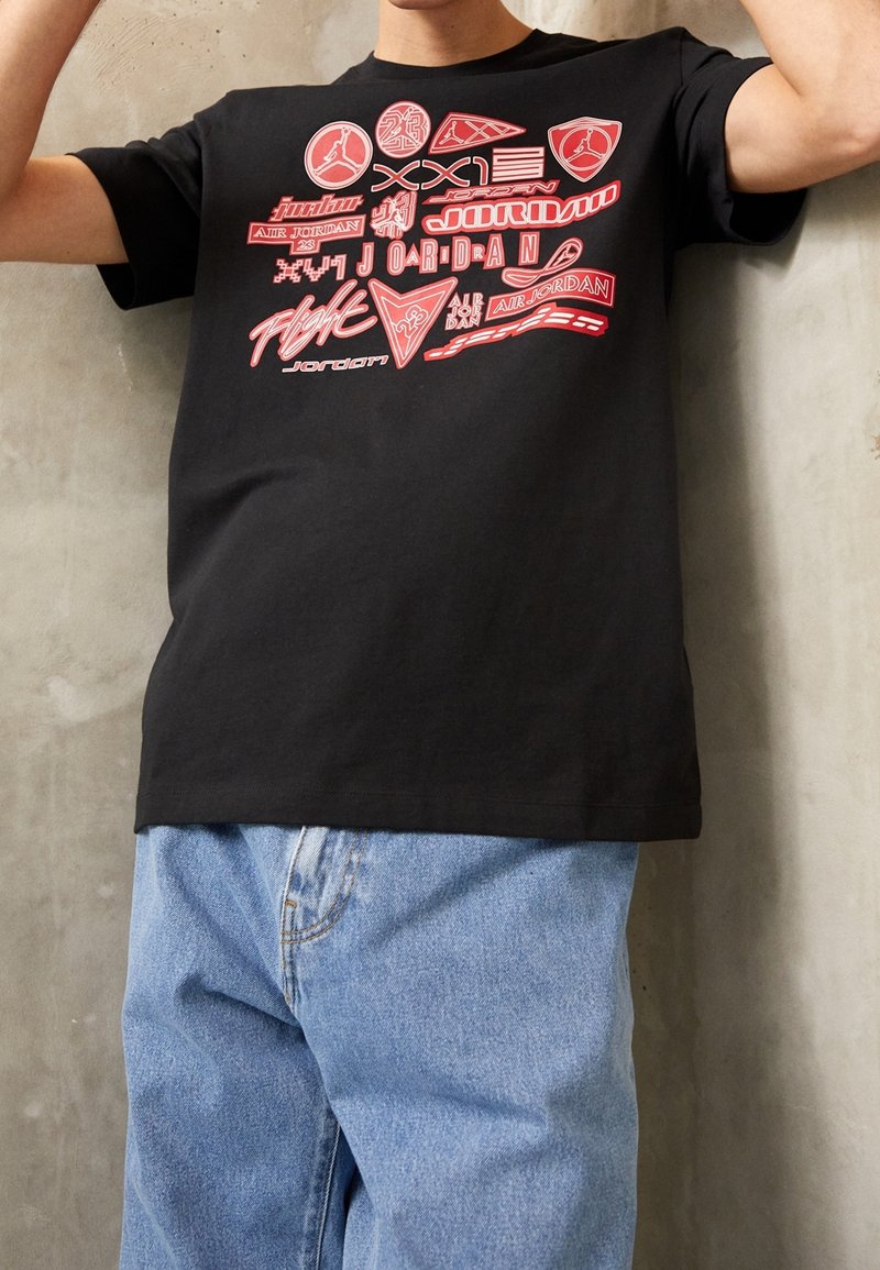 Camiseta negra con varios logotipos gráficos rojos y patrones de texto, de cuello redondo y mangas cortas, combinada con jeans de mezclilla sueltos de color azul.