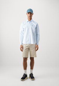 Lichtblauw gestreept overhemd met knopen, beige shorts, zwarte sneakers met beige zolen en een spijkeren pet. Eenvoudige en casual outfit.
