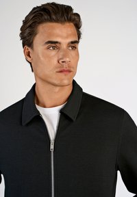 Zwarte jas met rits en kraag, gemaakt van gestructureerde stof. Opvallende zilveren ritssluiting. Draagt over een wit shirt.