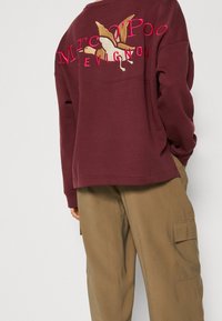 Bordeauxrotes Sweatshirt mit gestickten Motiven, darunter "Mr. Poo" und ein Vogeldesign, kombiniert mit locker sitzenden beigefarbenen Cargohosen.