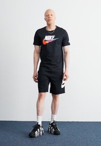 Črna Nike majica s pisanih logotipom, črne kratke hlače in črne superge z belimi poudarki. Be bele nogavice so vidne. Enostavna ozadje.