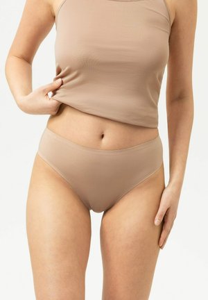Vrouw draagt beige naadloos ondergoed en tanktop, tilt het shirtje iets op om het middel te tonen tegen een effen achtergrond.