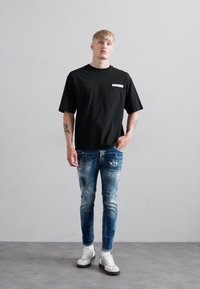 Jonge man met tatoeages, gekleed in een zwart "Obsession" t-shirt, versleten blauwe jeans en witte sneakers, staande tegen een effen grijze muur.