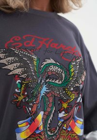 Grijze sweatshirt met een kleurrijk draakontwerp versierd met strasssteentjes, rode tekst en abstracte patronen in verschillende kleuren.