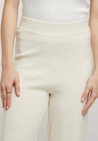Pantalons beiges taille haute en tissu doux, présentant une texture lisse et un design minimaliste. Mains posées sur les hanches, mettant en valeur la taille.