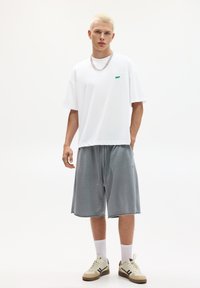 Witte oversized t-shirt met een groen logo, gecombineerd met grijze shorts met trekkoord, witte sokken en beige sneakers met donkere accenten.