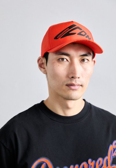 Rød baseballcaps med svart logo, rund skygge og strukturert design. Bært med en svart t-skjorte prydet med livlig blå tekst.