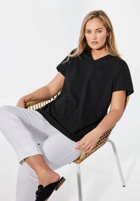 Schwarzes V-Ausschnitt-T-Shirt mit kurzen Ärmeln, aus glattem Stoff, kombiniert mit weißen, schmal geschnittenen Hosen und schwarzen Slippers, sitzt auf einem Korbstuhl.