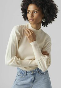 Maglione a maniche lunghe color crema con colletto alto e polsini a coste, abbinato a jeans a vita alta blu chiaro. Texture liscia.