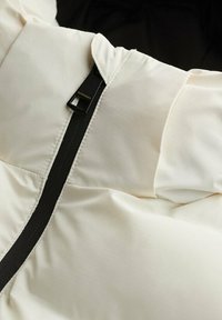 Acercamiento a una chaqueta acolchada de color crema con un tirador de cremallera negro etiquetado "Superdry" y detalles visibles de costura.