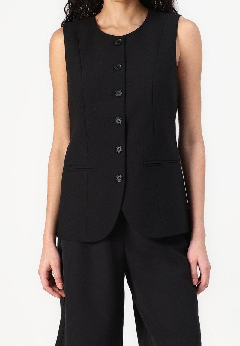 Femme portant un gilet noir sans manches boutonné et un pantalon large noir assorti sur un fond uni.