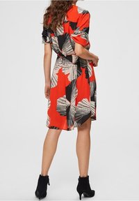 Vestito rosso con motivo di foglie nere e bianche, maniche corte, vita con cintura, lunghezza al ginocchio, tessuto liscio, abbinato a stivaletti neri alla caviglia.