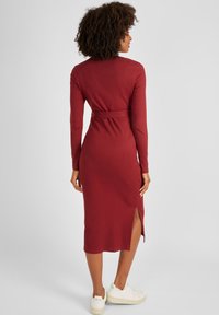 Langärmliges, geripptes Kleid in dunklem Rot mit tailliertem Schnitt und seitlichem Schlitz, kombiniert mit weißen Sneakern. Glatte Textur, knielanges Design.