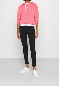 Kvinna klädd i en rosa huvtröja med vita snören, svarta slim-fit jeans och vita sneakers, som står mot en enkel ljusgrå bakgrund.