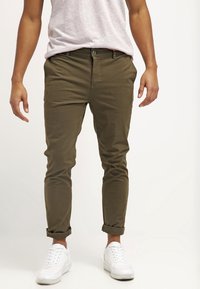 Pantaloni verde oliva in cotone, dal taglio aderente, con risvolti alle caviglie e tasche standard, abbinati a sneakers bianche.