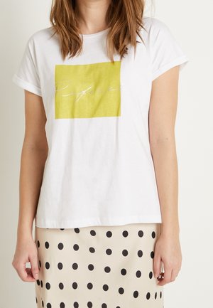 Hvid bomulds-T-shirt med optrukne ærmer, der har et grønt firkantet grafisk tryk med sølvskrift. Bæret med en prikket nederdel.