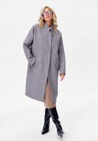 Manteau en laine gris surdimensionné avec un col haut, des manches larges et des poches latérales, superposé sur une robe beige et associé à des bottes noires jusqu'aux genoux.