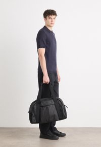 Μαύρη τσάντα σάκος με διπλές λαβές, θήκες με φερμουάρ και ανάγλυφο λογότυπο. Δομημένος σχεδιασμός με ομαλή υφή και ανθεκτικό υλικό.