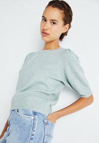 Pull en maille vert clair avec de courtes manches bouffantes et un ourlet côtelé, associé à un jean en denim taille haute. Texture lisse avec une coupe décontractée.