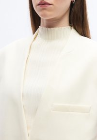 Γυναίκα που φοράει εκρού ριμπ πλεκτό ζιβάγκο κάτω από ένα εκρού ραμμένο σακάκι με τσέπη στο στήθος και μικρά σκουλαρίκια κρίκους.