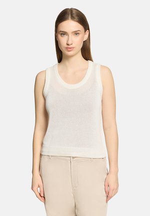 Jeune femme aux cheveux bruns raides, portant un top en maille blanc sans manches et un pantalon beige, debout devant un fond clair uni.