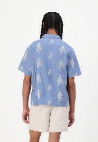Chemise bleue à manches courtes et col, ornée d'un motif blanc en forme de corail, fabriquée en tissu léger, associée à un short beige.