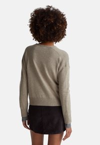 Kobieta z kręconymi włosami, ubrana w beżowy sweter z długim rękawem i ciemną spódnicę, widziana od tyłu na jednolitym tle.